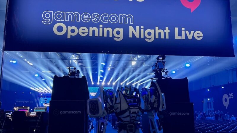 A robot mascot standing on stage beneath the "Welcome to gamescom Opening Night Live" banner in a blue-lit arena before the 2025 show.