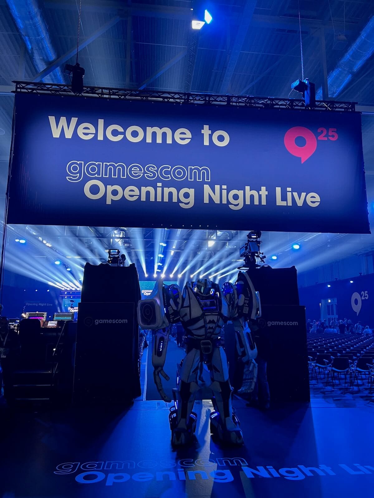 A robot mascot standing on stage beneath the "Welcome to gamescom Opening Night Live" banner in a blue-lit arena before the 2025 show.